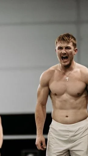 A shirtless man with short hair and a cross necklace, yelling or shouting, in an indoor setting.