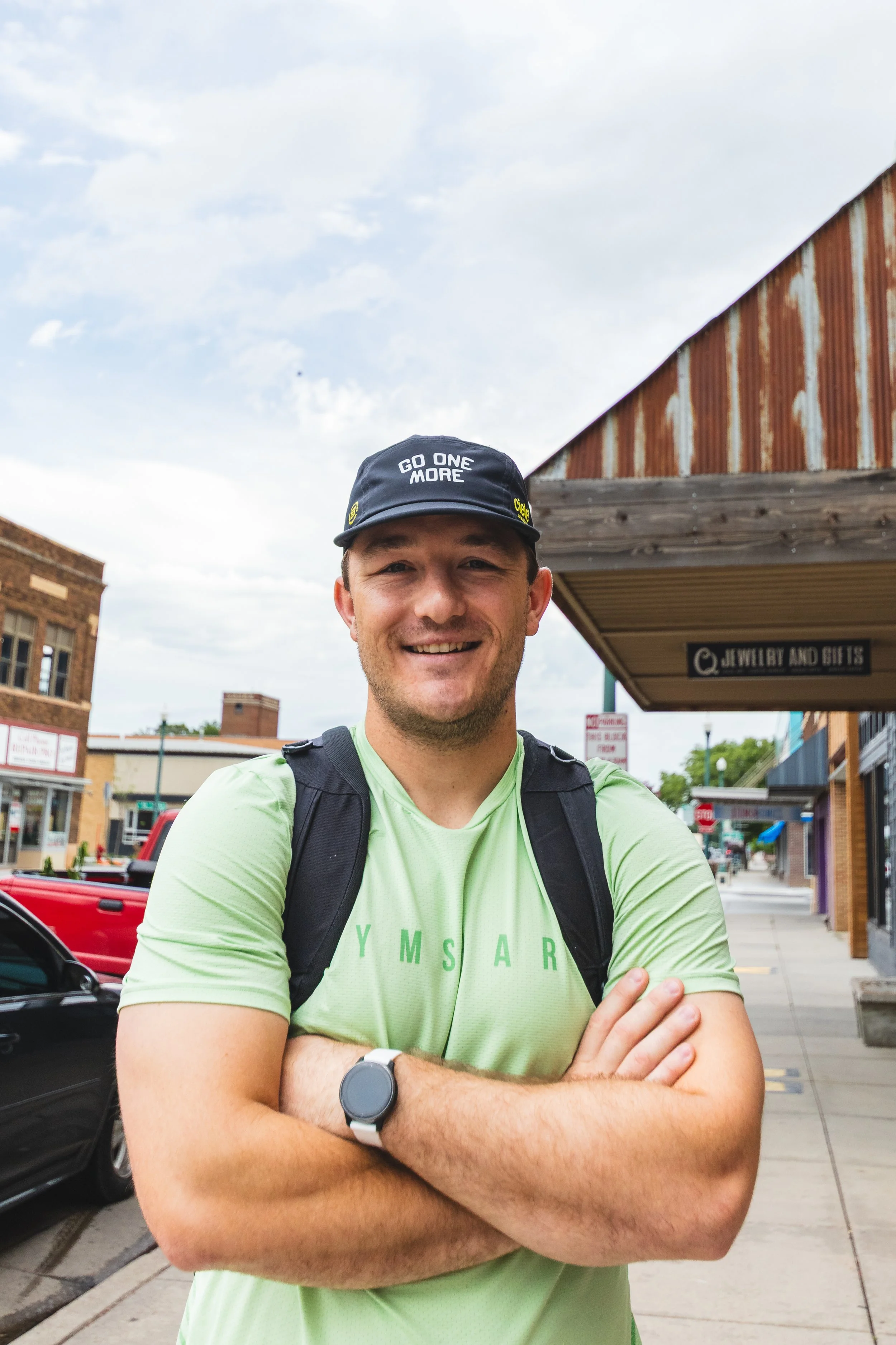 A man with a backpack smiling and crossing his arms on a city sidewalk, wearing a baseball cap that says 'Go One More' and a lime green T-shirt, with storefronts and buildings in the background.