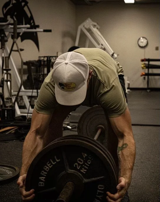 Man in a beige t-shirt and baseball cap lifting a barbell in a gym.