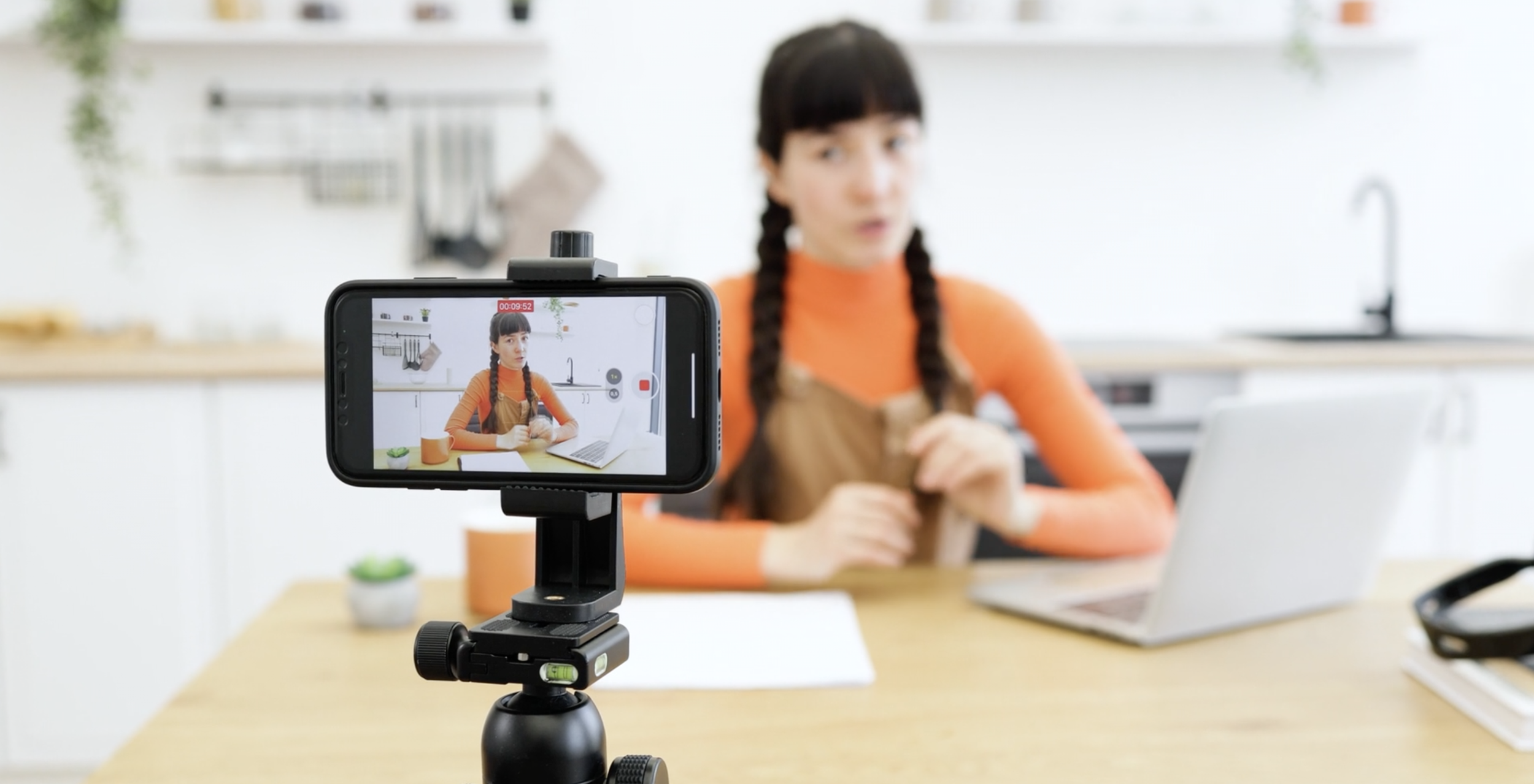 A woman with dark hair in braids sits at a kitchen table, filming herself with a smartphone mounted on a tripod. The kitchen has white cabinets, a sink, and modern decor.