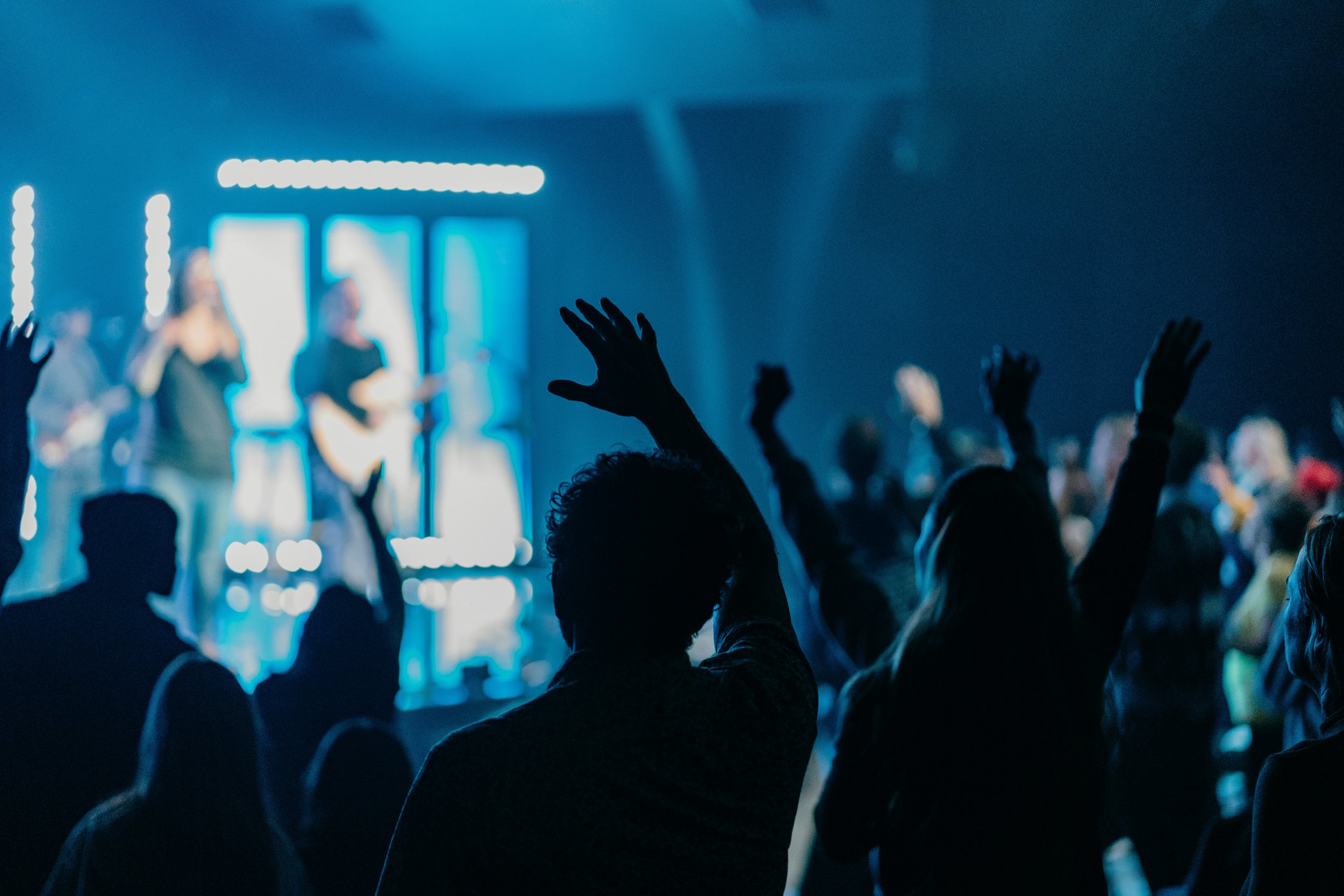 People with raised hands at a concert or event, with a blurred stage and performers in the background.