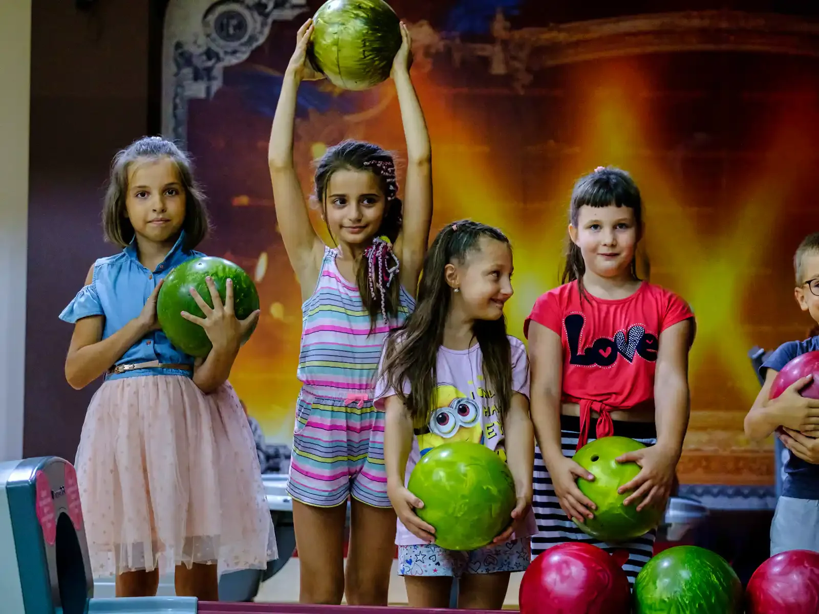 Group of young children holding colorful bowling balls indoors, with a decorated background.