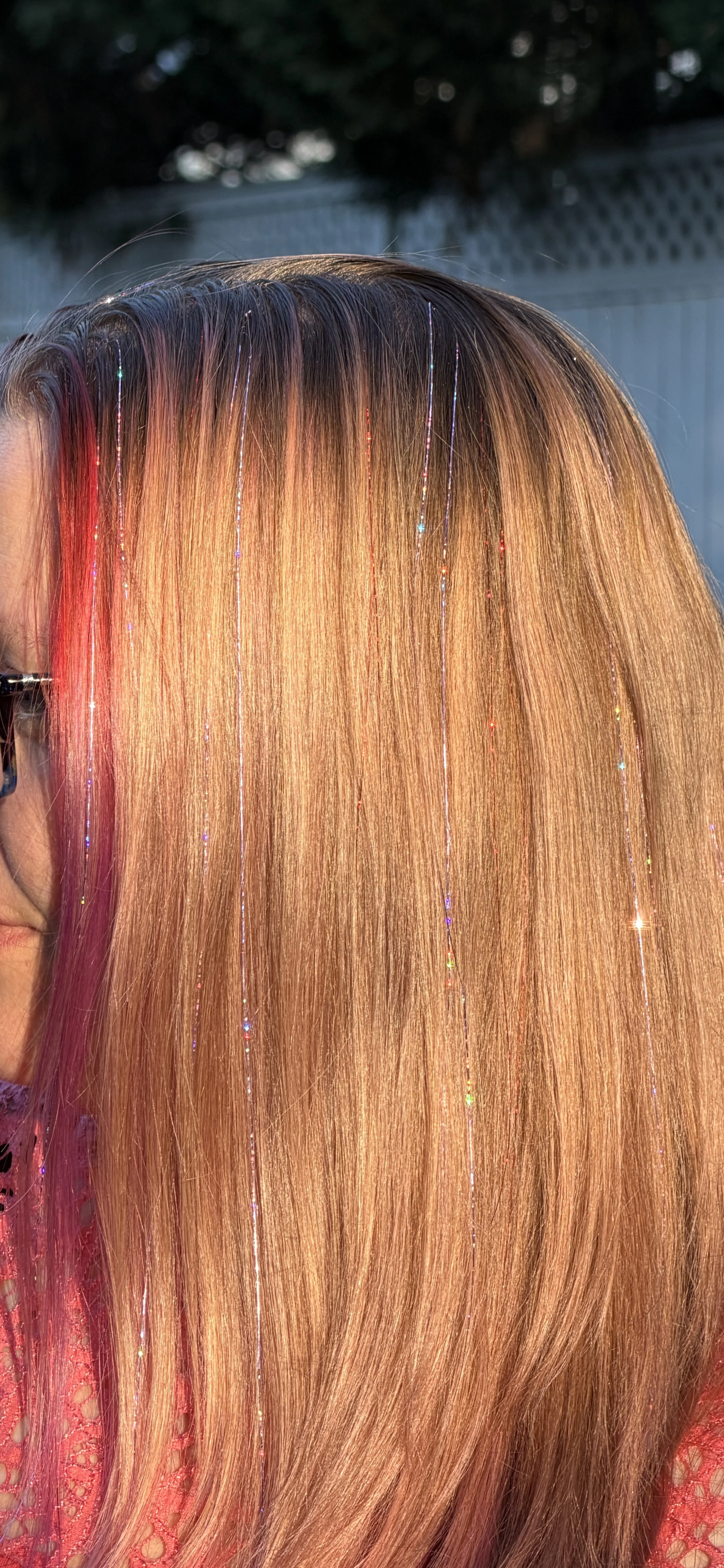 Close-up of long, straight, shiny hair with pink and blond hues, decorated with thin, colorful, reflective strands, outdoors with a blurred background.