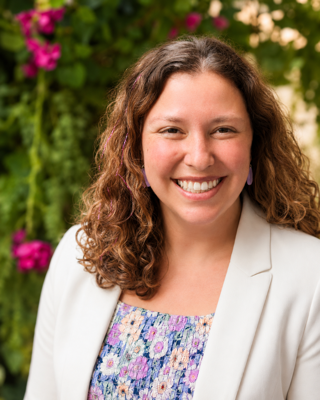 A woman with curly brown hair smiling outdoors with pink flowers and green foliage in the background, wearing a white blazer and a colorful floral top.