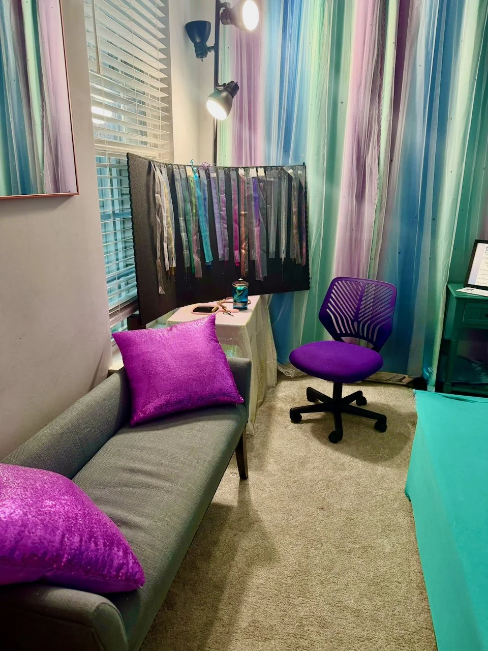 A room with a gray sofa and purple pillows, a purple office chair, a small teal-colored desk, and a black board with various ribbons hanging on it. The room has colorful curtains with pastel shades, a window with white blinds, and overhead lighting.