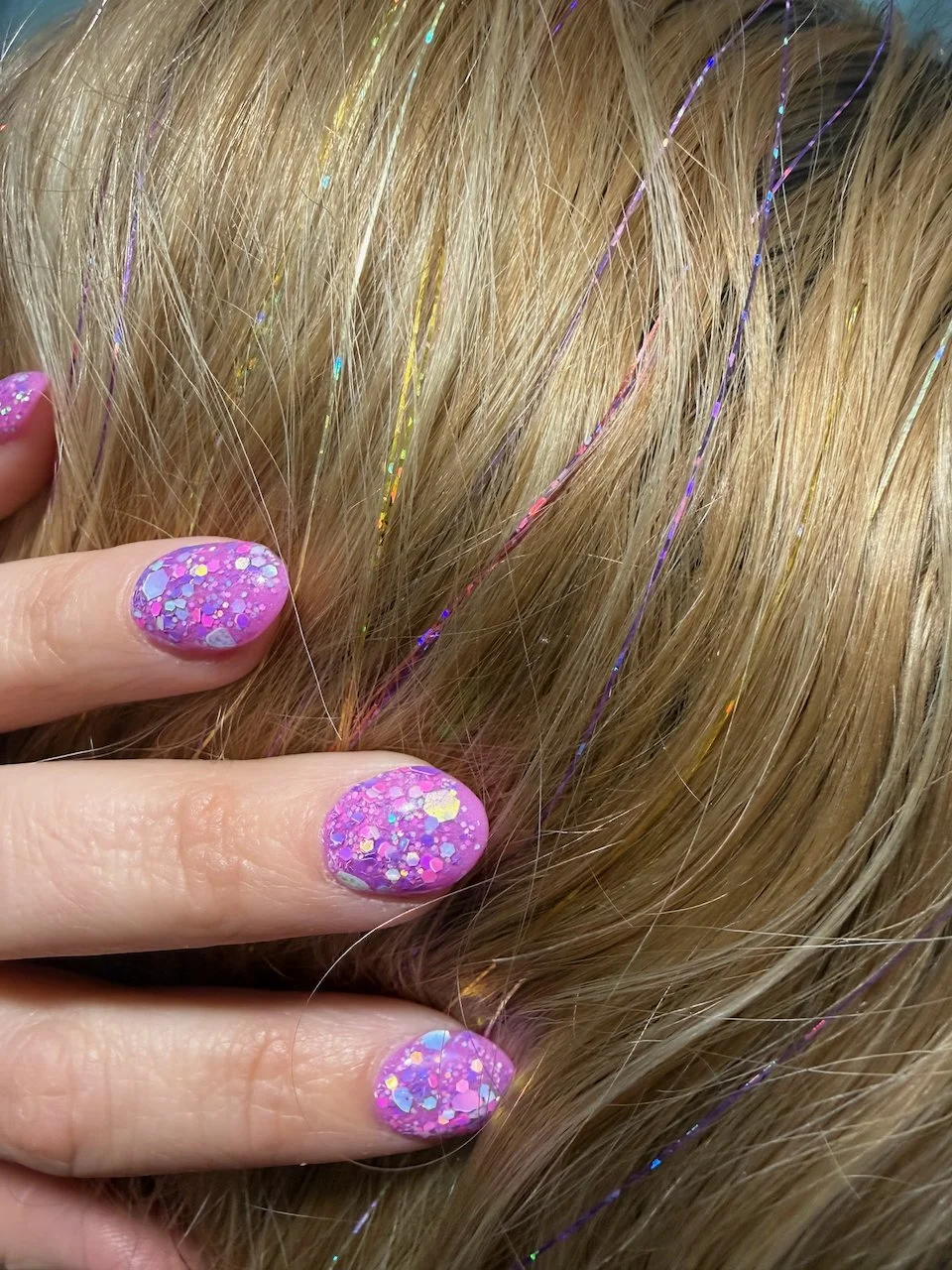 Close-up of blonde hair with colorful tinsel and pink nails with glitter, fingers holding strands of hair.