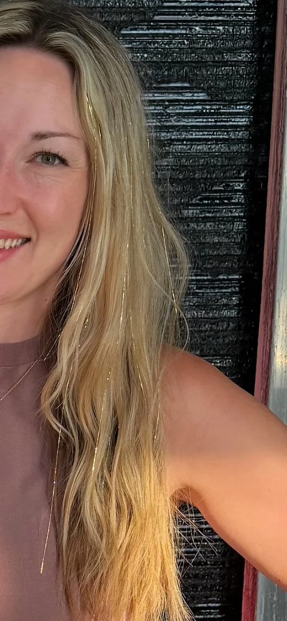 Close-up of a smiling woman with long wavy blonde hair, wearing a sleeveless top, standing outdoors near wooden and metal structure.