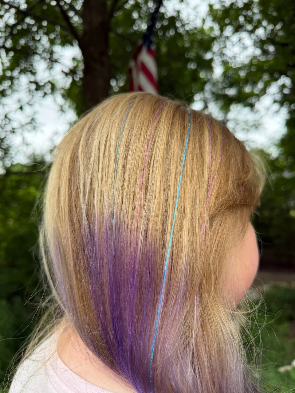 Child with blonde hair, dyed purple at the ends, with thin multicolored streaks of blue, pink, and purple, outdoors with leafy trees and an American flag in the background.