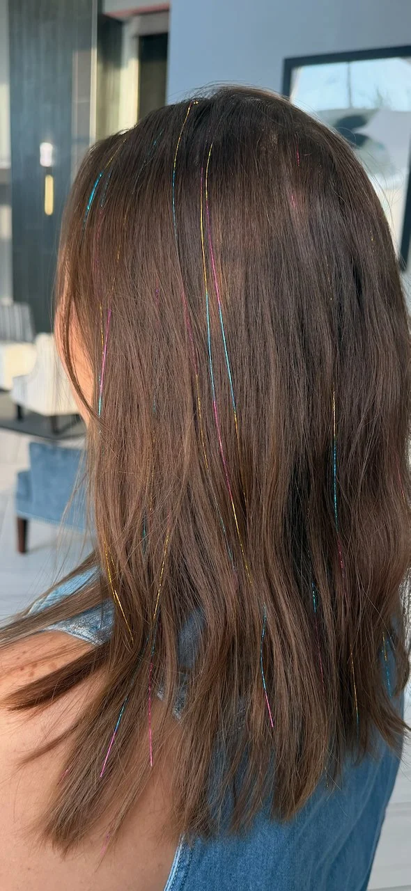 Person with brown hair and colorful, thin, metallic hair strands woven throughout, sitting indoors.