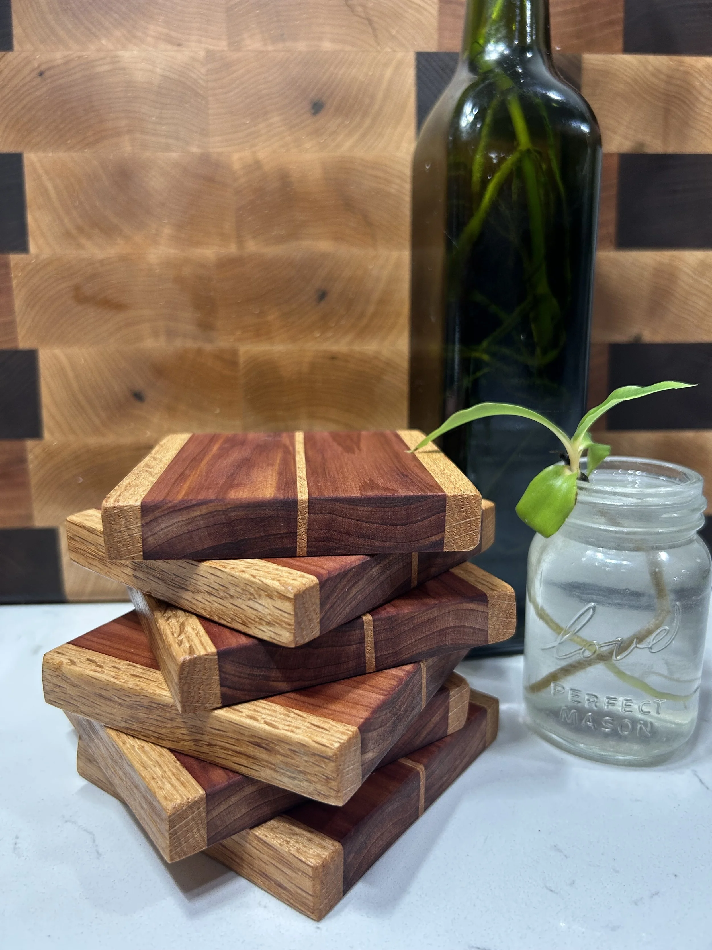 Stacked wooden coasters in front of a small glass jar with a plant inside and a dark green vase containing tall green plant stems on a wooden surface.
