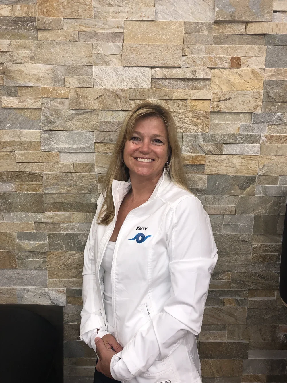 A woman with blonde hair smiling, wearing a white jacket with a logo and the name 'Kerry' on it, standing in front of a textured stone wall.