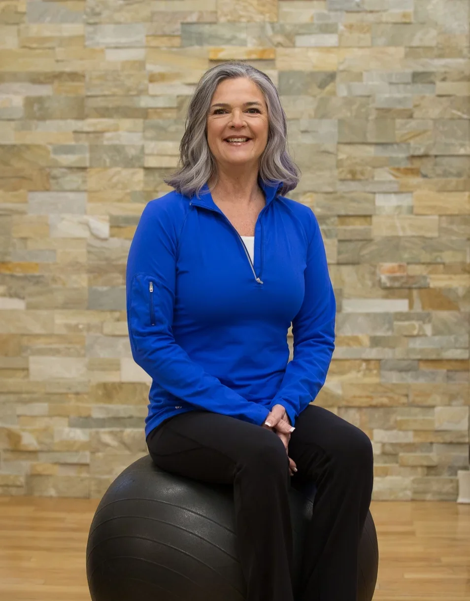 A smiling middle-aged woman with gray hair sitting on a black exercise ball in a gym or fitness studio, wearing a blue athletic jacket and black pants, in front of a beige stone wall.