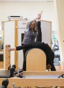 A woman practicing Pilates in a gym, bending over with one leg raised and another foot on the Pilates equipment, with a trainer assisting her.