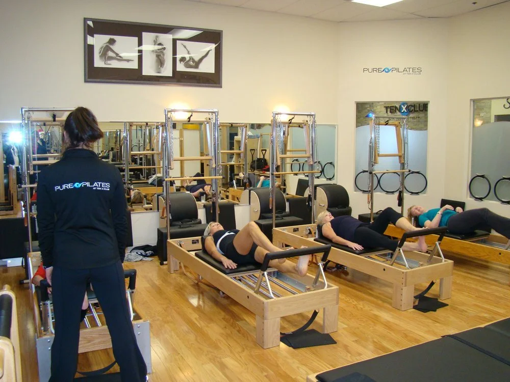 Pilates class in a studio with participants lying on reformer machines, instructor supervising, mirrors, and fitness equipment.