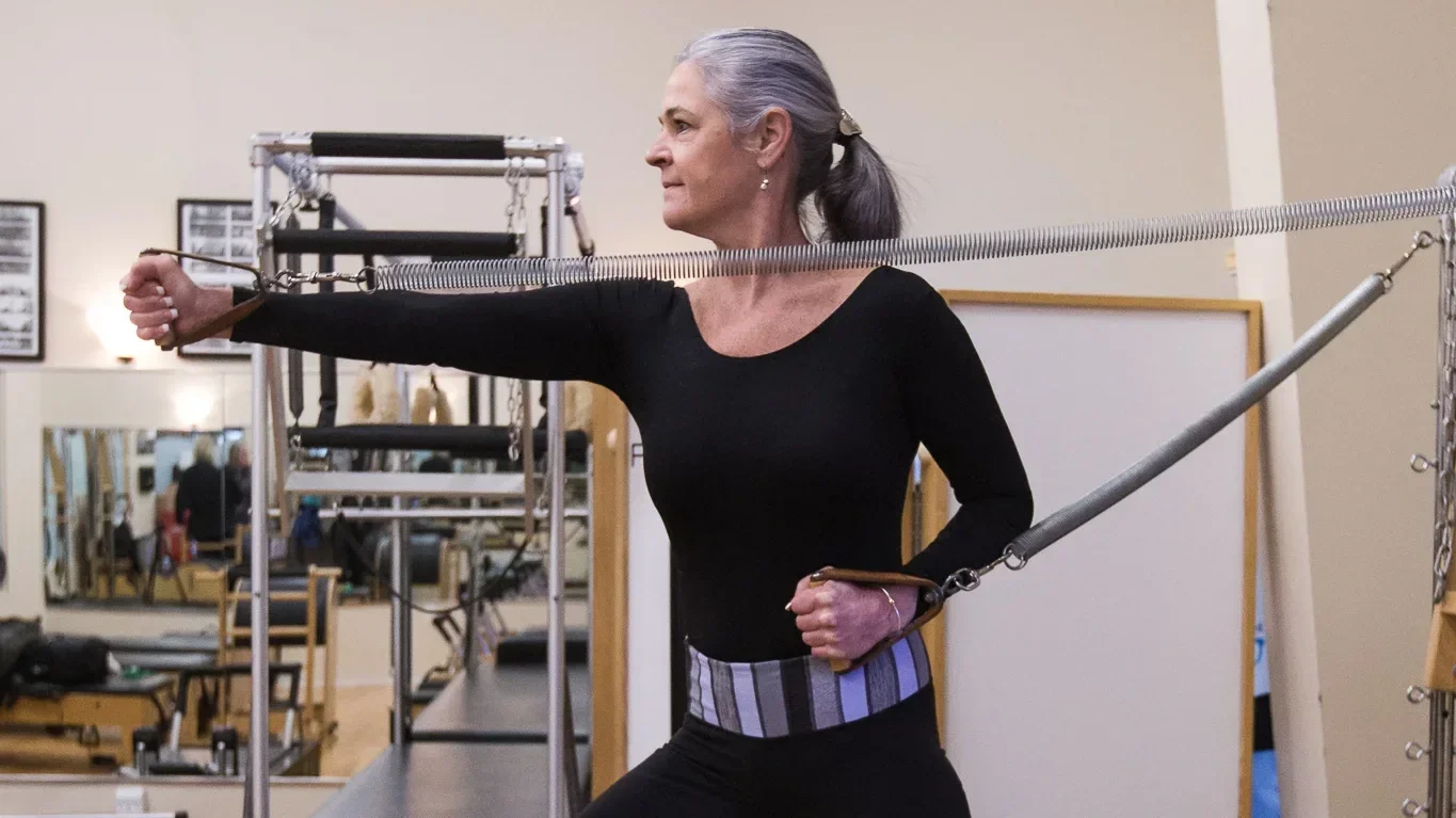 An older woman with gray hair tied back in a ponytail exercising on a Pilates reformer machine in a gym, wearing a black long-sleeve top and a striped support belt around her waist.