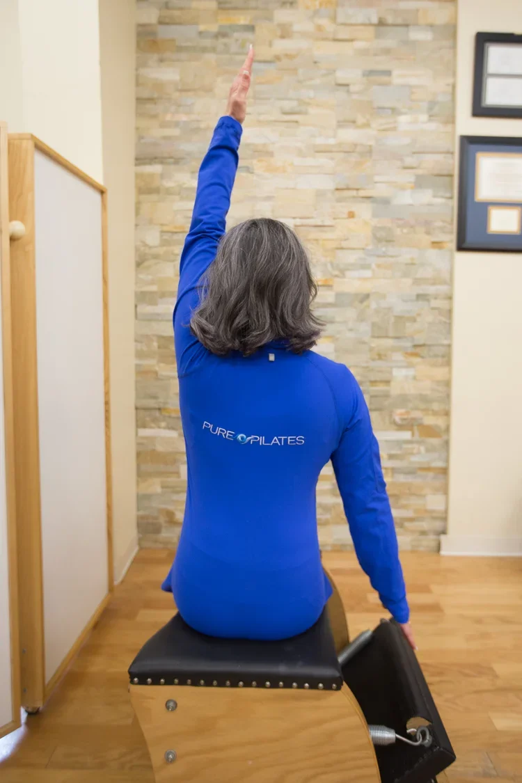 A woman in a blue 'Pure Pilates' shirt sitting on a Pilates reformer machine, raising her right arm overhead in a stretching or exercise position, in a room with a stone accent wall and framed certificates.