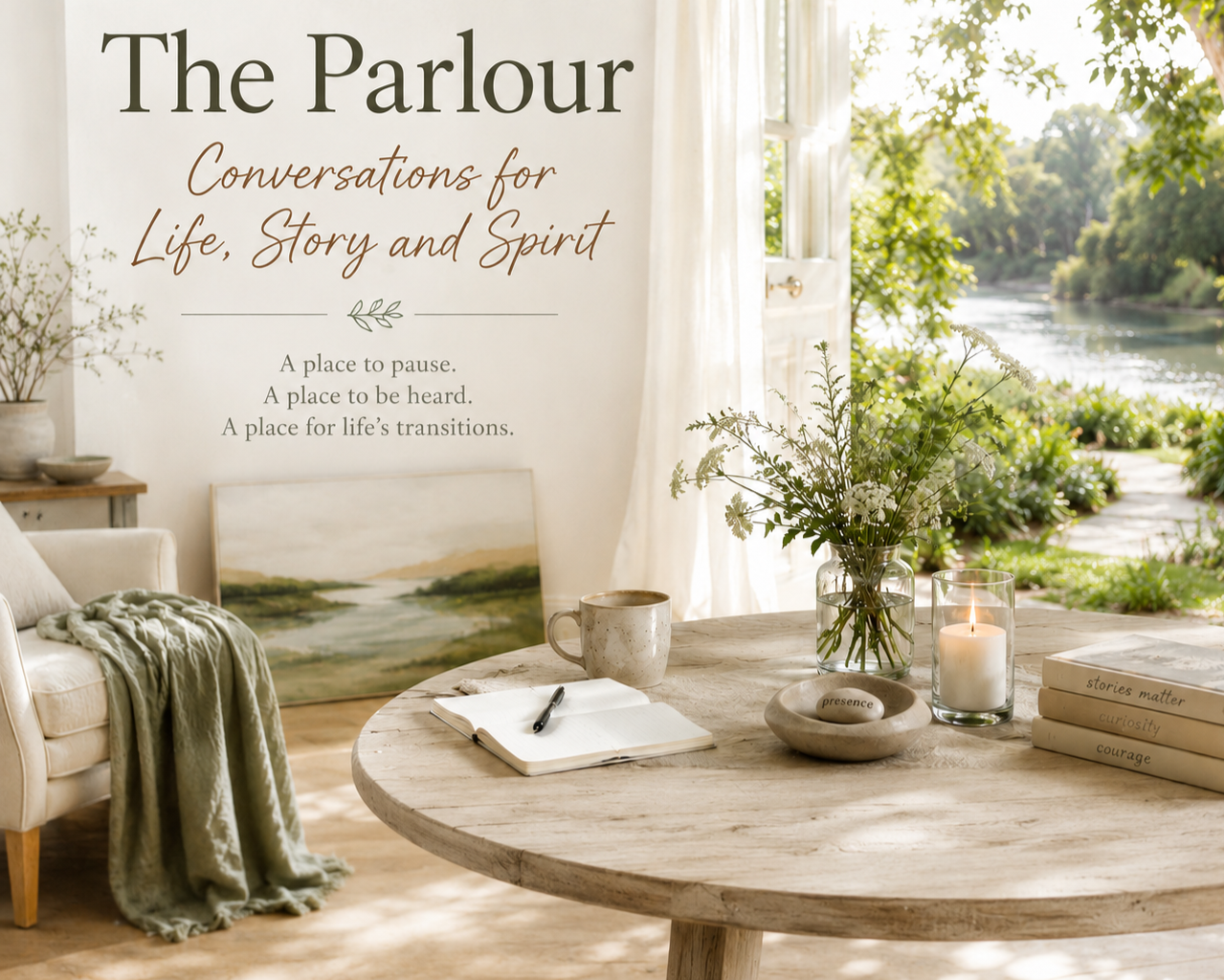 A cozy interior with a round wooden table holding a notebook, pen, mug, vase with greenery, candle, and three books titled 'stories matter,' 'curiosity,' and 'courage.' In the background, a large window shows a river and trees. A wall displays text about 'The Parlour' and its purpose.