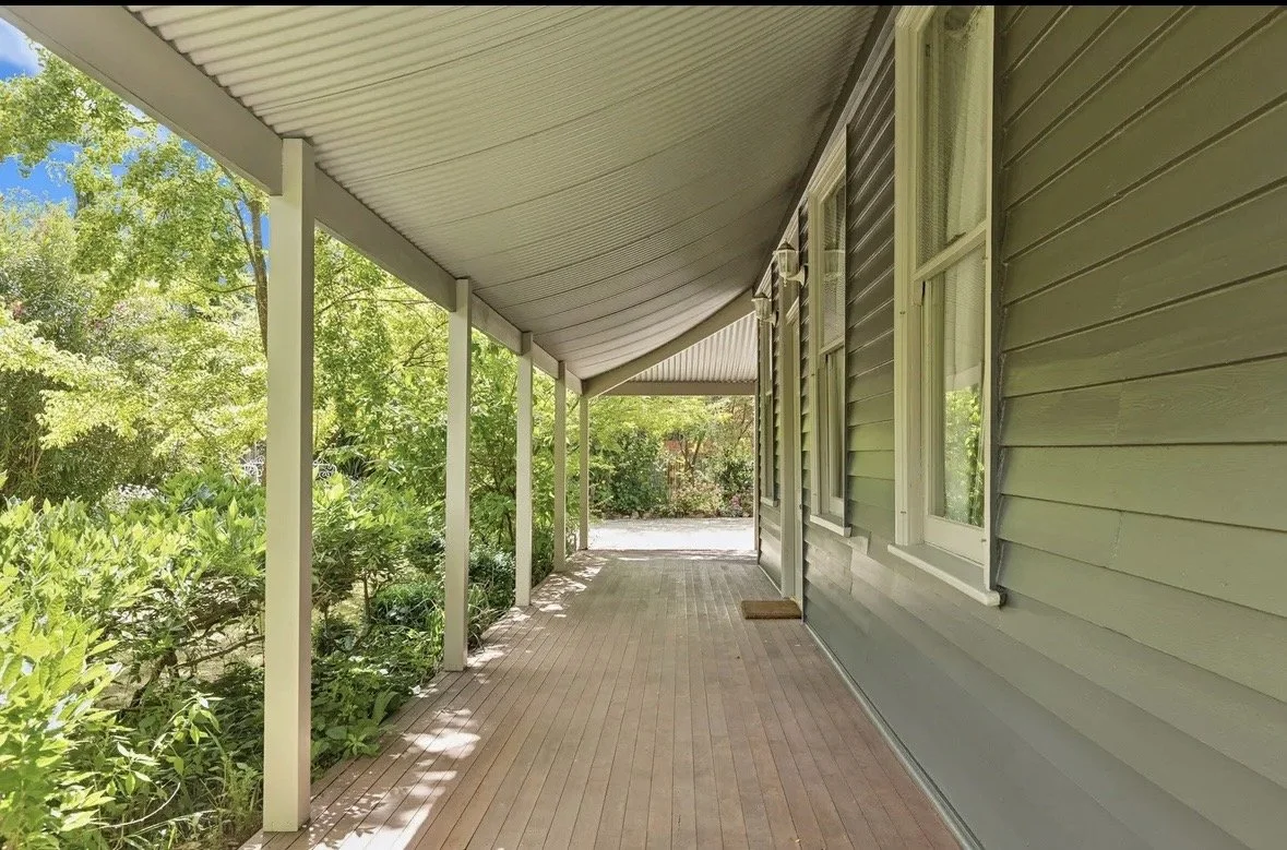 Covered porch with gray siding house exterior and green foliage in the background.