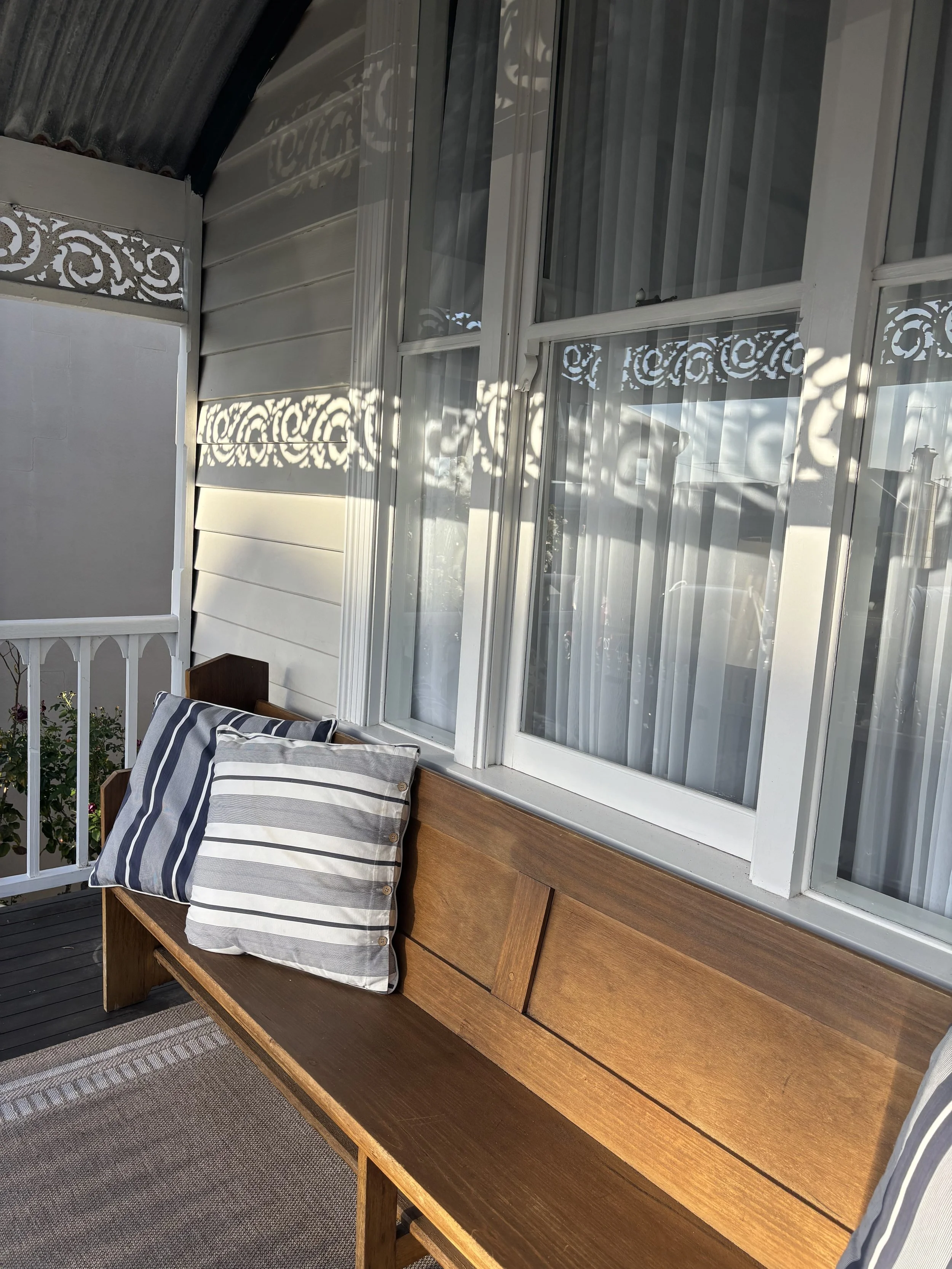 A wooden bench with a striped pillow placed on it on a porch with large windows decorated with white lace curtains and a decorative white railing casting shadows.