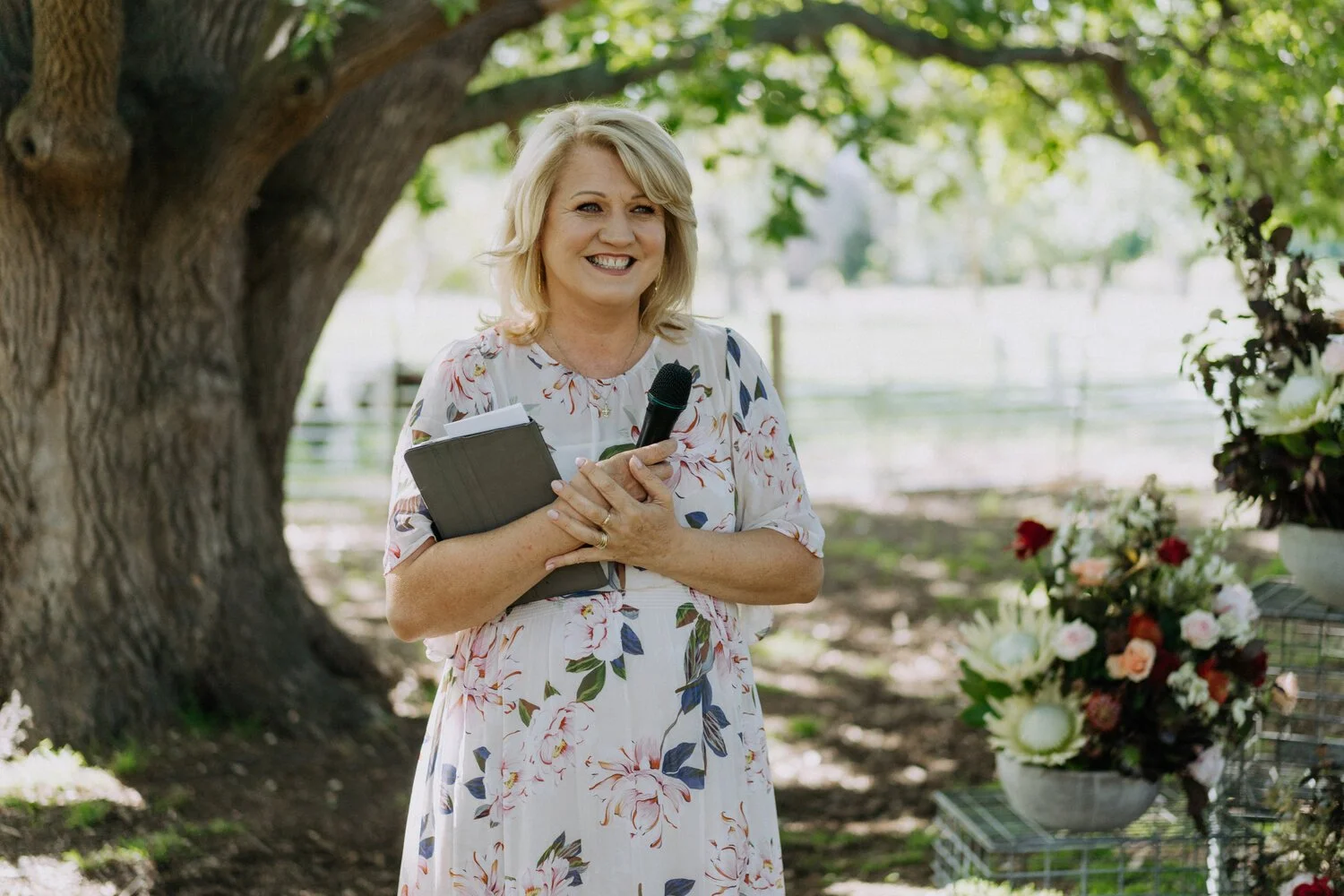A woman with blonde hair, wearing a floral dress, is smiling while holding a microphone and a notebook outdoors under a large tree, with flower arrangements nearby.