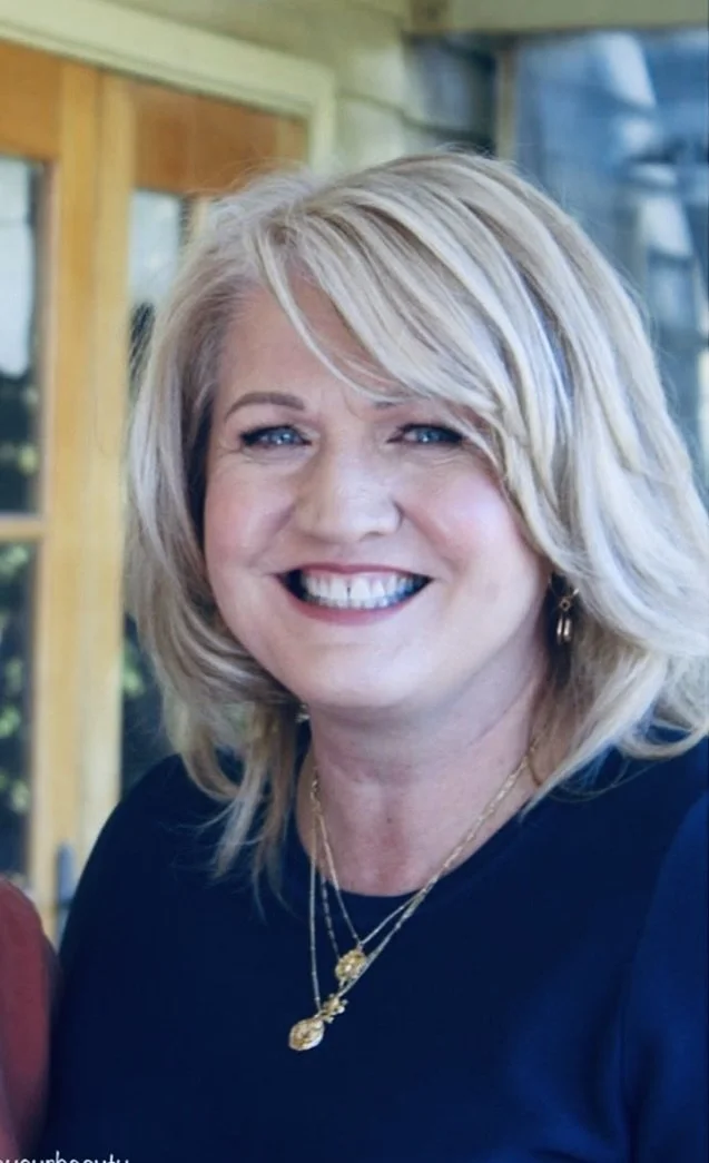 A smiling woman with shoulder-length blonde hair, blue eyes, wearing a dark top, gold necklace, and earrings, standing indoors with windows and green surroundings visible in the background.