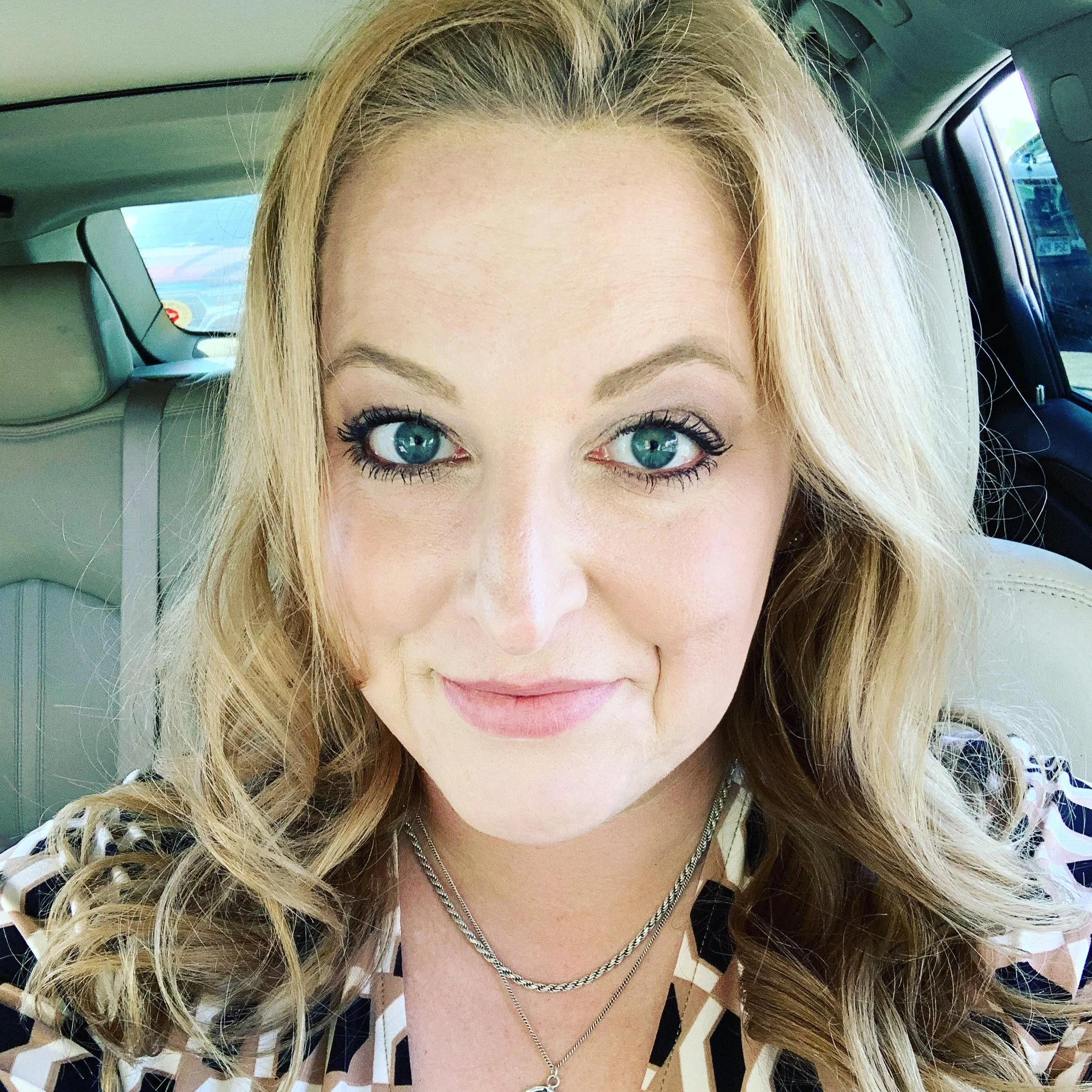 Close-up of a woman with blonde hair and blue eyes, sitting in a car with beige interior