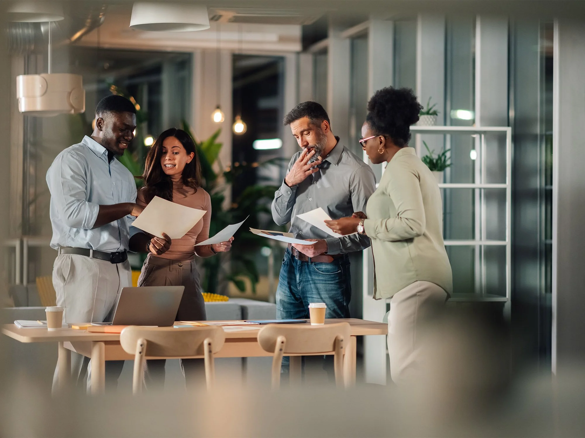 Four diverse professionals studying documents around a table in a modern office with large windows, hanging lights, and indoor plants.