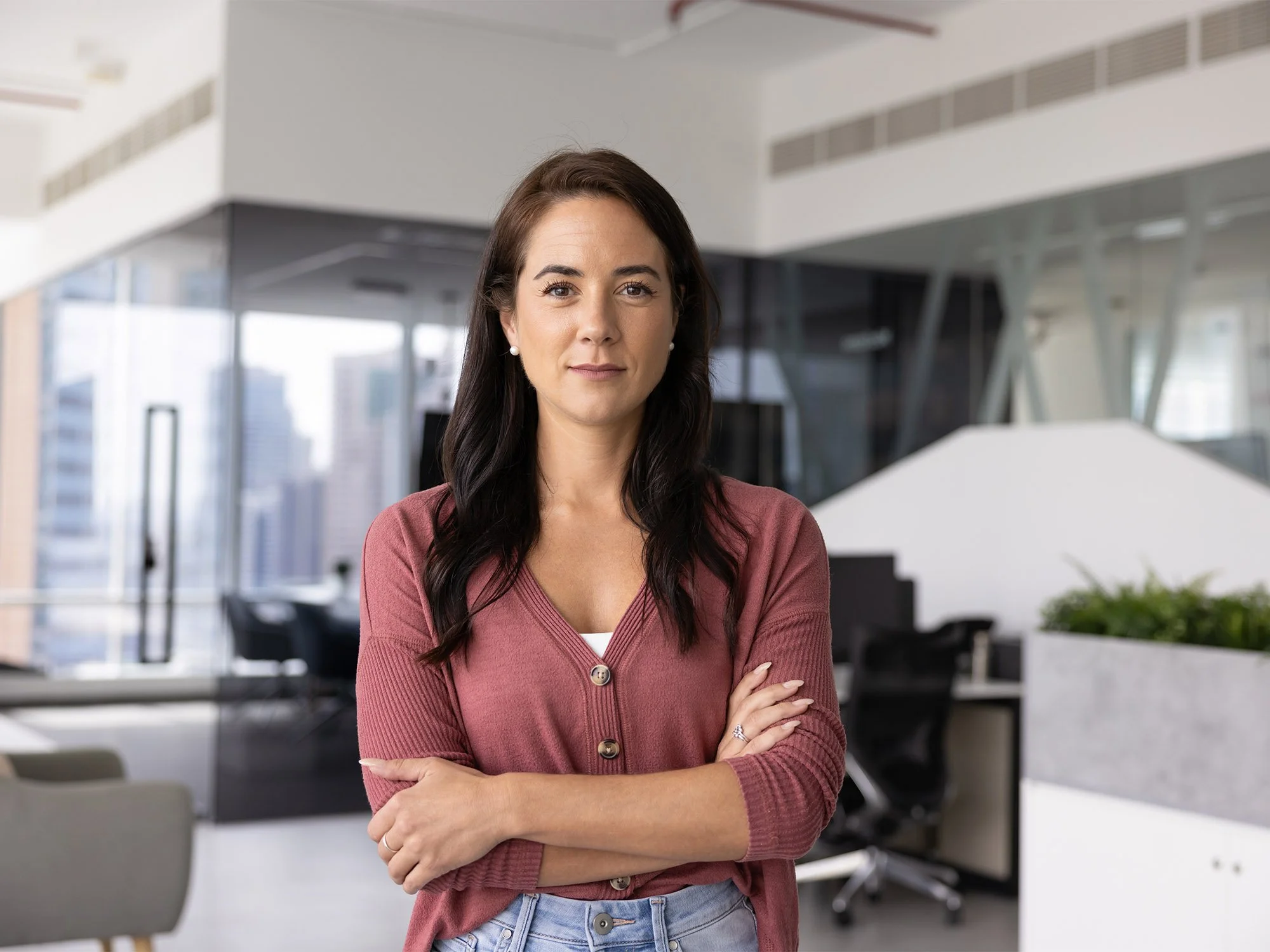 A woman with dark brown hair, wearing a maroon cardigan, stands confidently with arms crossed in a modern office space with large windows and urban city view.