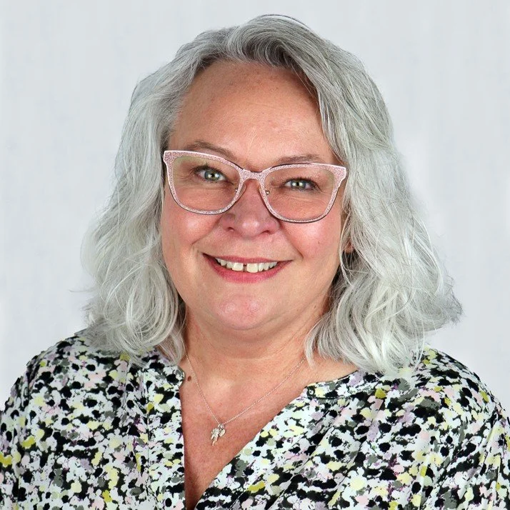 A woman with gray, curly hair wearing pink glasses and a floral blouse, smiling in front of a plain background.
