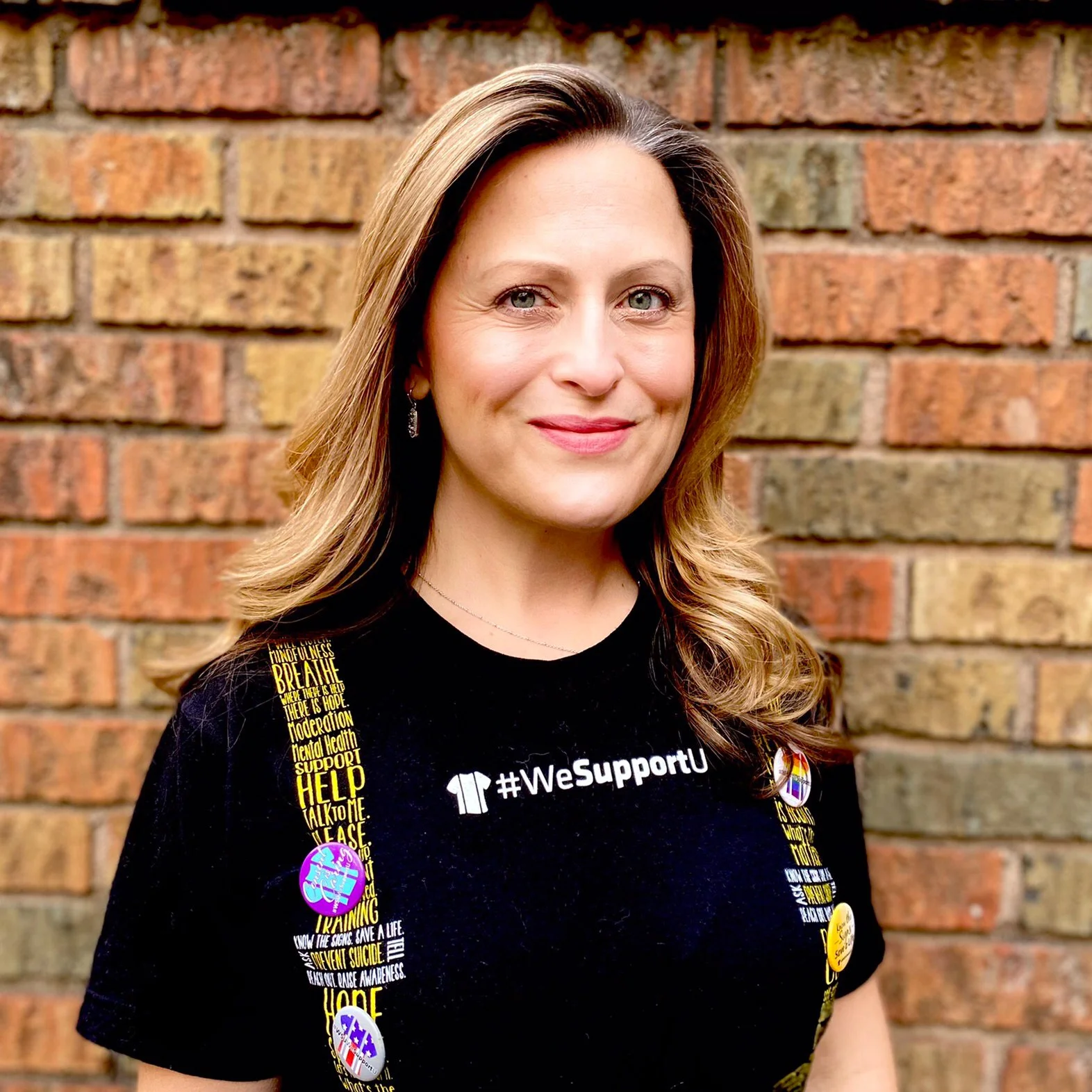 A woman with long, blonde hair, wearing a black t-shirt with #WeSupportU printed on it, standing outdoors in front of a brick wall, smiling at the camera.