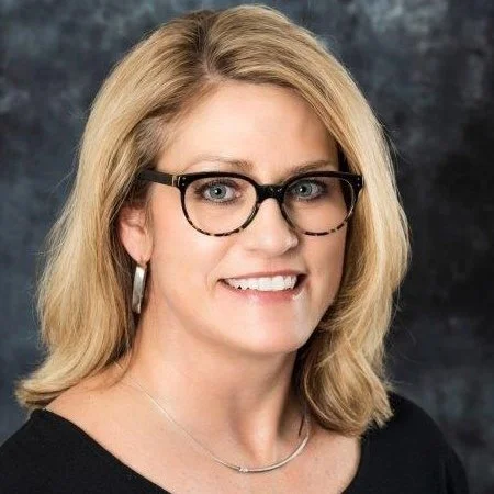 A woman with blonde hair and glasses smiling against a dark background.
