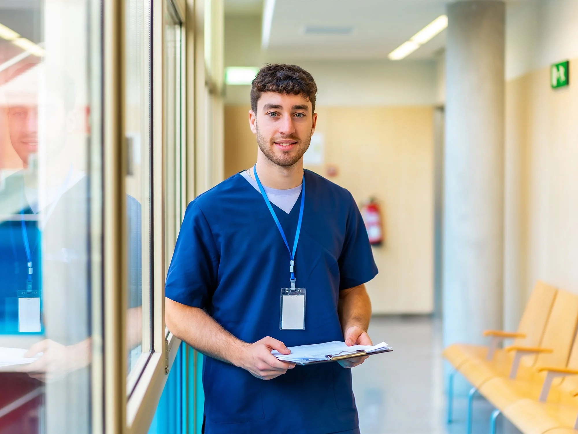 Young male nurse in blue scrubs with a lanyard around his neck holding a clipboard, standing in a hospital hallway with windows and yellow chairs.