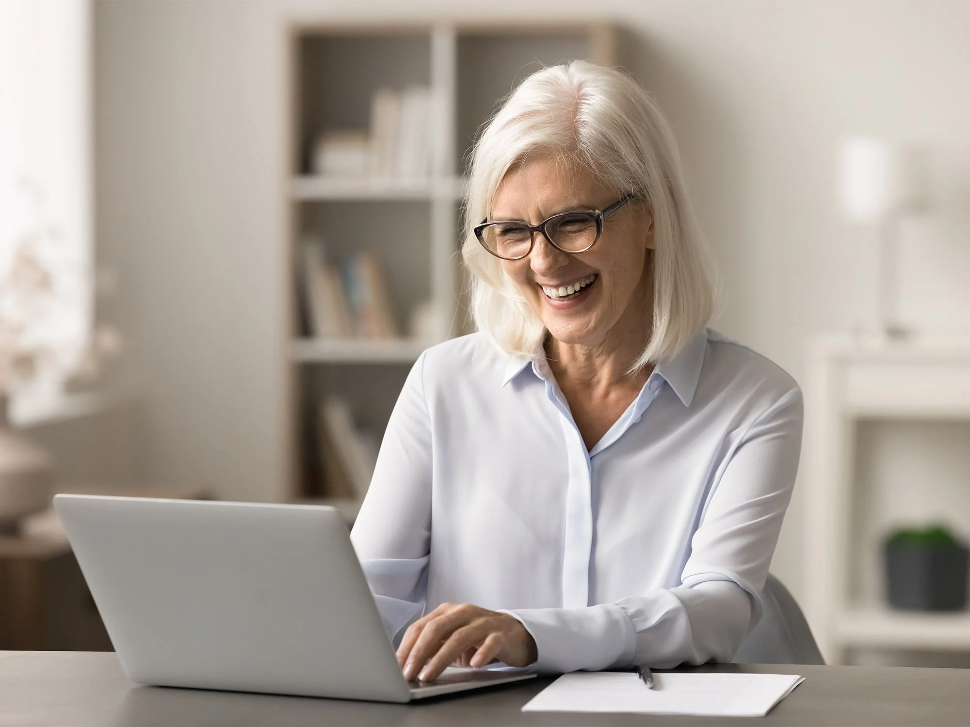 An older woman with white hair and glasses working on a laptop in an office setting, smiling while looking at the screen.