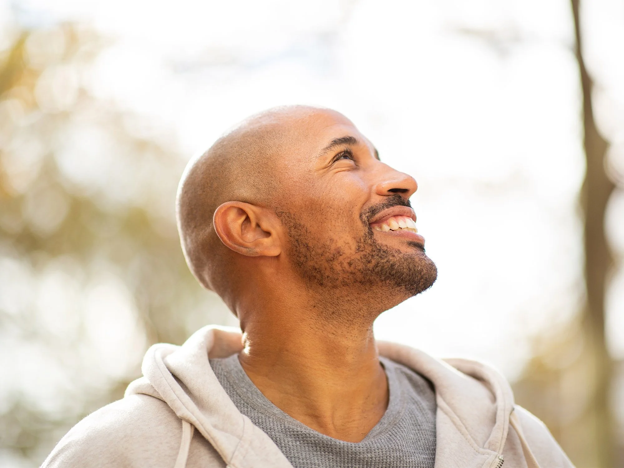 A smiling man with a shaved head and beard outdoors, looking up and to the right, with a blurred background of trees and sunlight.