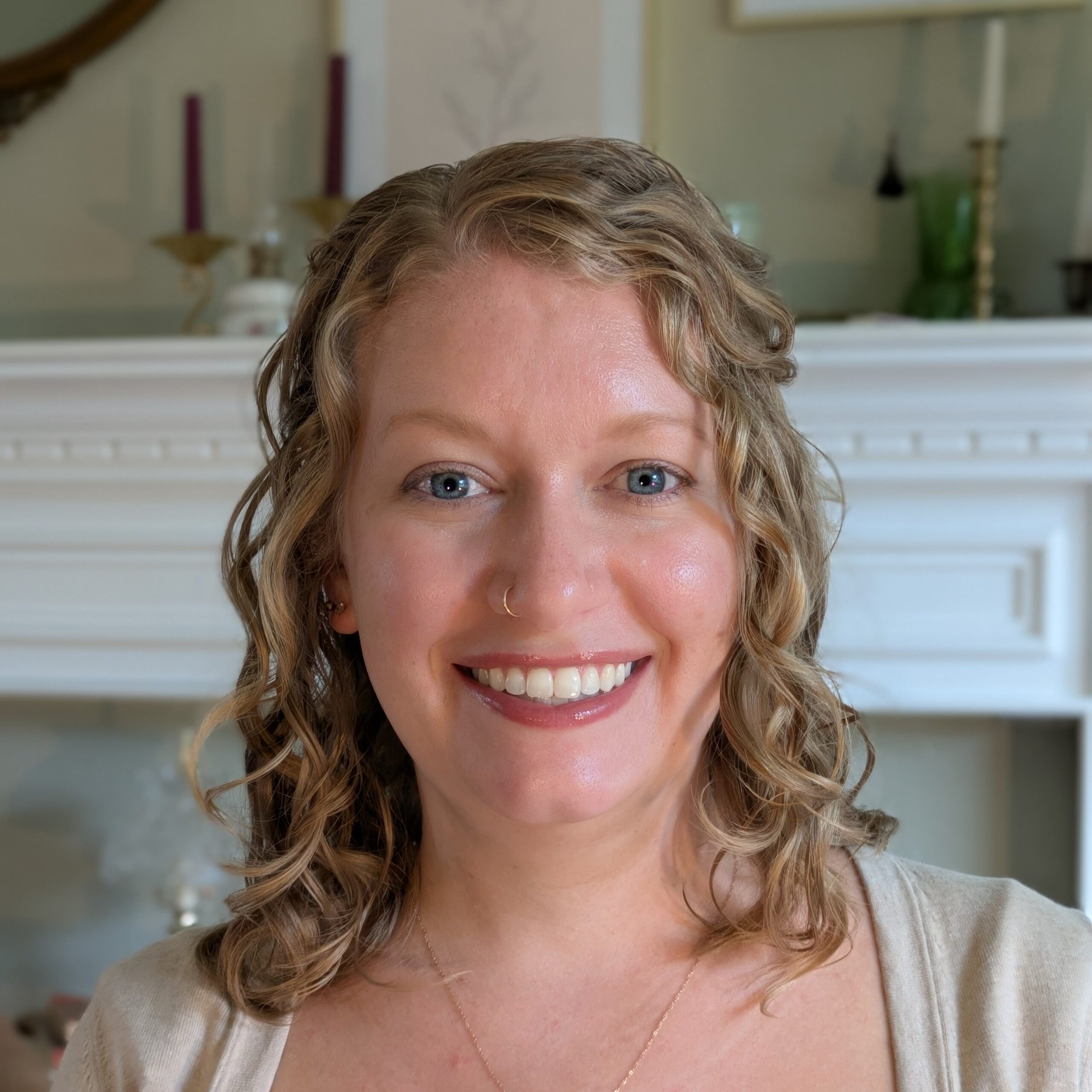 A woman with blonde, curly hair and blue eyes smiling, wearing a light-colored top and a delicate necklace, with a white fireplace and decorative items in the background.