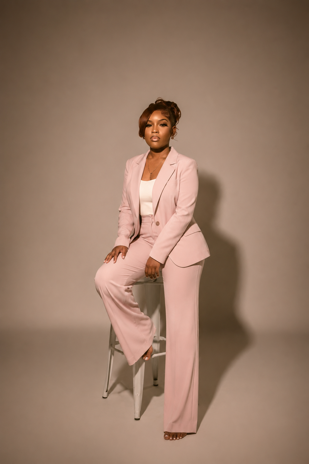 A woman dressed in a light pink suit, sitting on a stool against a plain background.