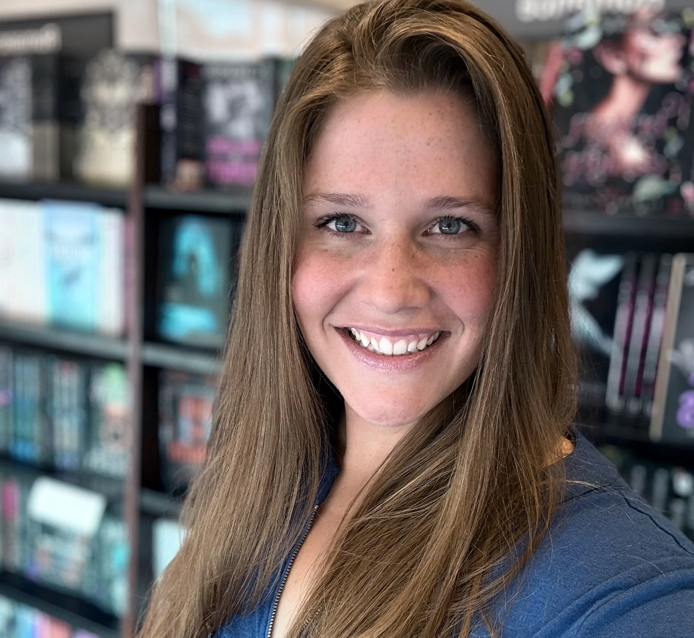 Close-up of a smiling woman with long, light brown hair and blue eyes, standing in front of a bookshelf filled with books.