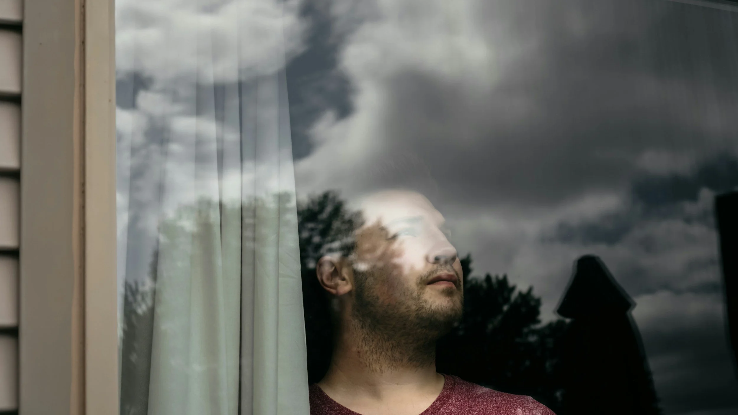 A man with closed eyes looking out a window with cloudy skies and trees reflected on the glass.