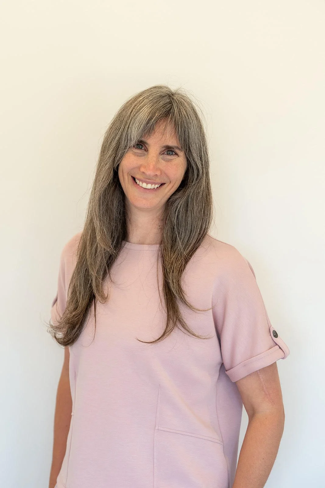 Smiling woman with long brown hair wearing a light pink t-shirt standing against a plain white wall.