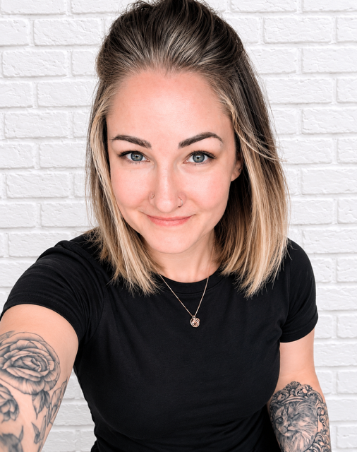 Woman with blonde hair, wearing a black t-shirt, standing in front of a white brick wall, smiling at the camera.