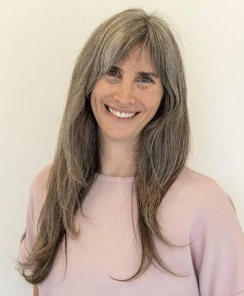 A woman with long gray hair wearing a light pink top smiling in front of a plain wall.