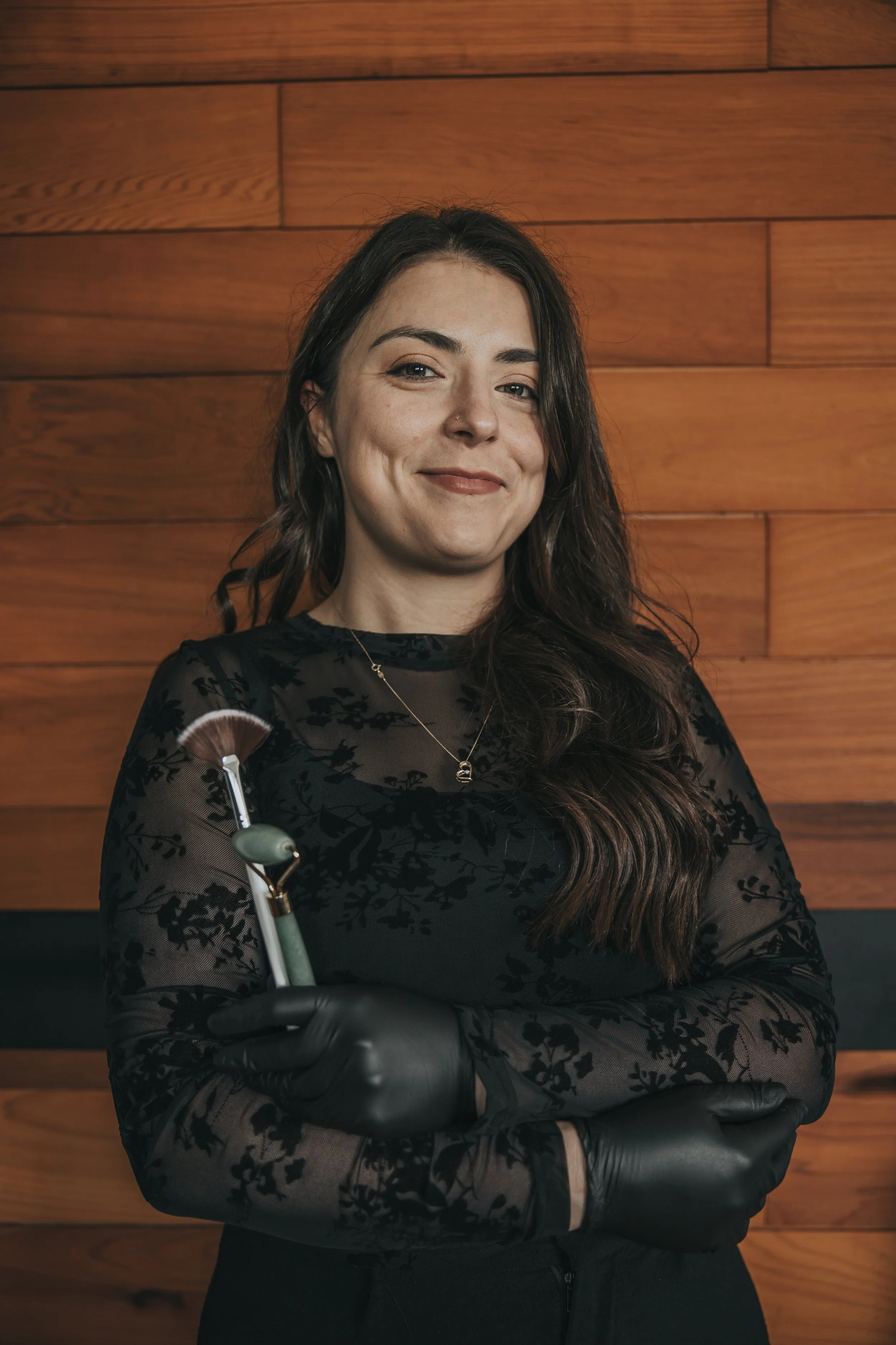 Woman smiling, holding a dental tool, wearing black gloves, against a wooden wall background.