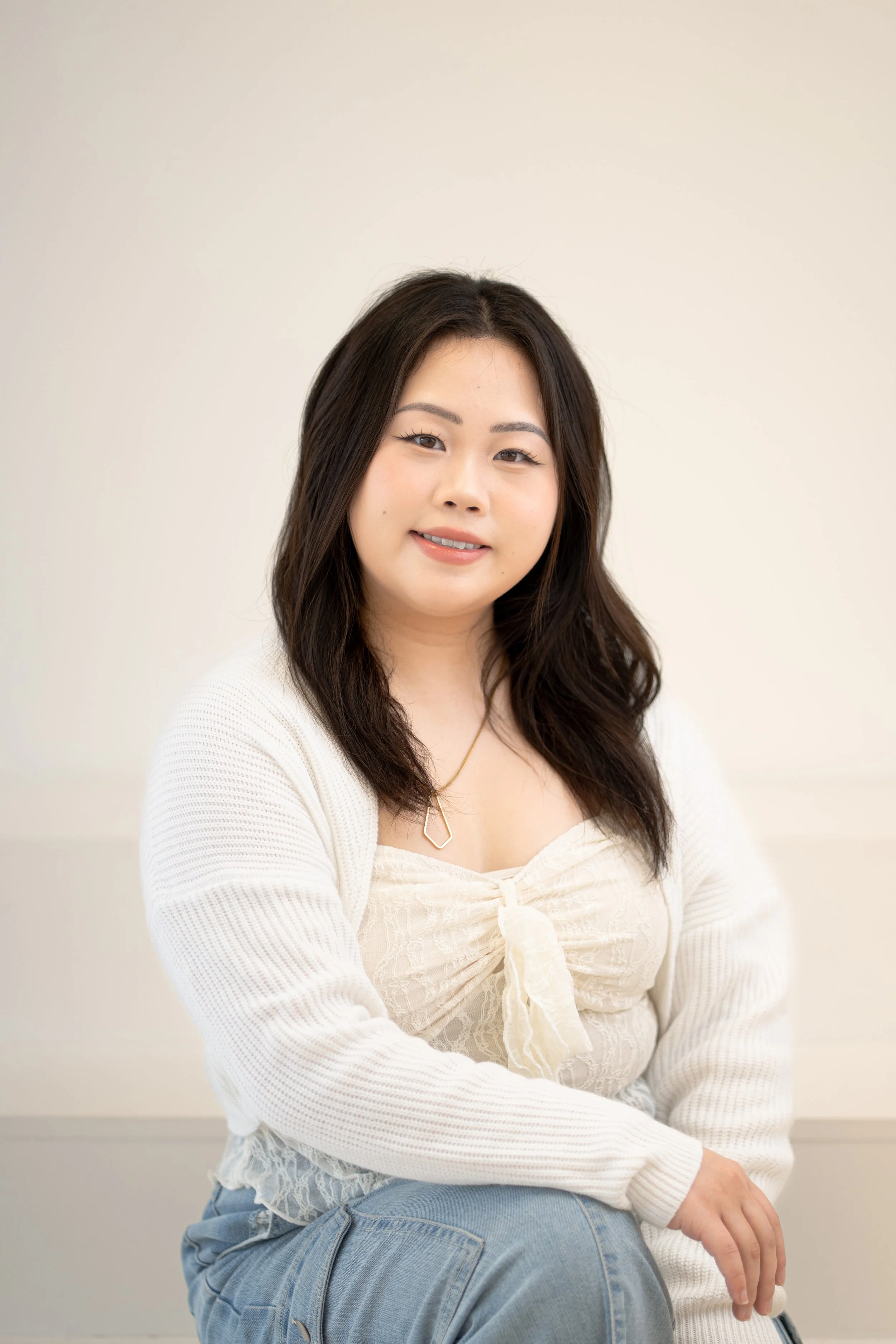 A young woman with long dark hair sitting against a plain light-colored wall. She is smiling slightly and wearing a cream-colored blouse with lace details and a bow, a white cardigan, and blue jeans.