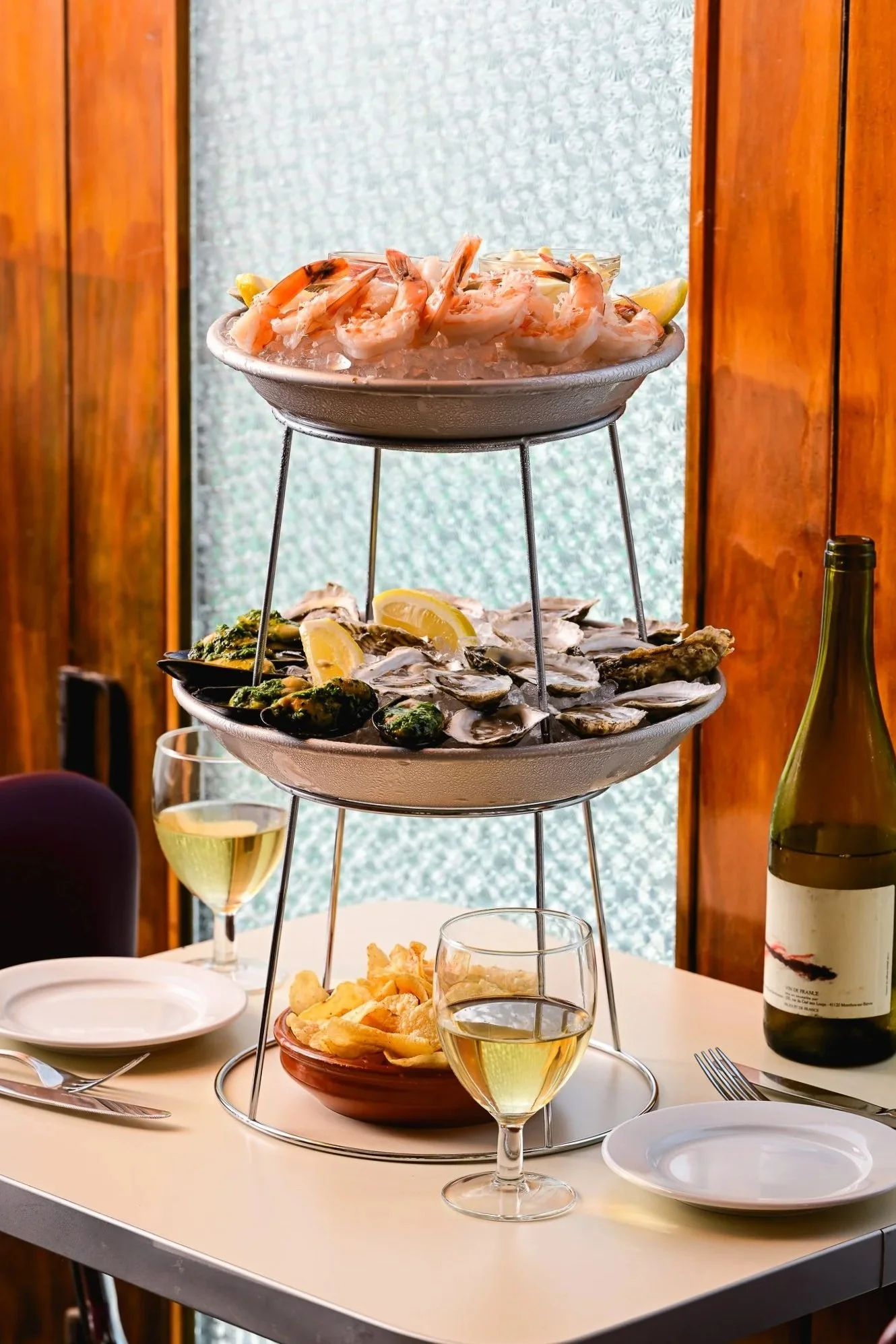 A three-tiered seafood platter with shrimp on the top, oysters on the middle, and mussels with lemon wedges on the bottom. The platter is set on a white table with wine glasses, a bottle of white wine, and side dishes.