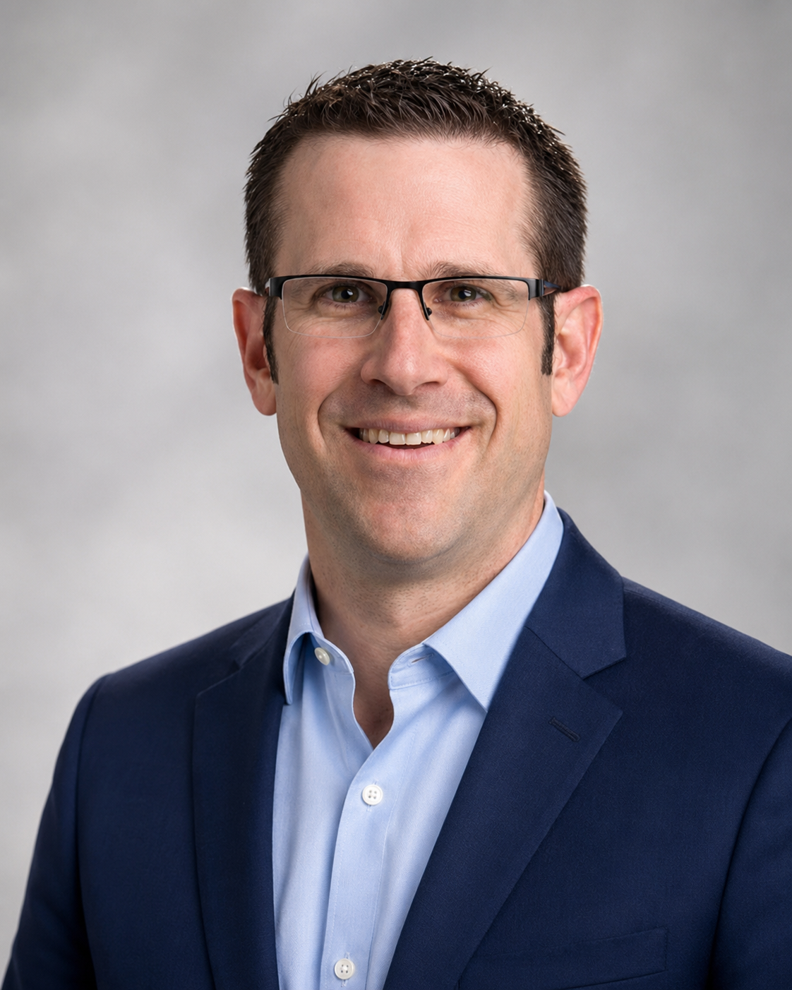 Professional headshot of a man wearing glasses, a blue suit, and a light blue shirt, smiling against a neutral background.