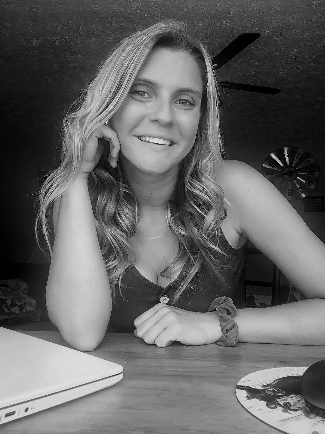 A young woman with curly hair smiling and resting her head on her hand while sitting at a desk with a closed laptop and a mouse pad.