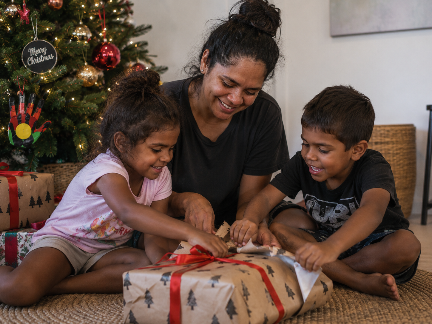 A woman and two children opening a Christmas gift under a decorated Christmas tree.