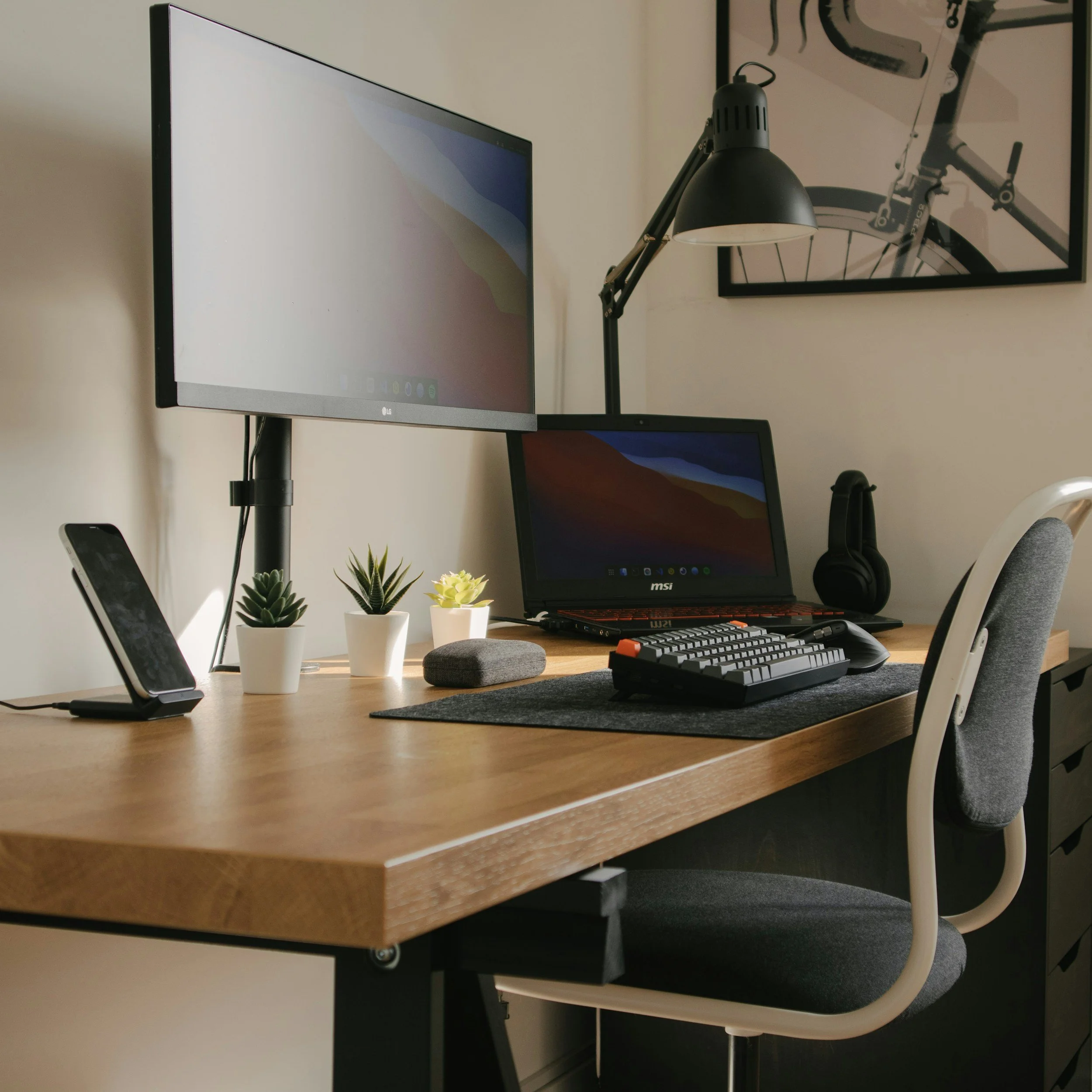 A workspace with a desk, a large monitor, a laptop, two small potted plants, a keyboard, and a computer mouse. There is a desk lamp and a framed picture of a bicycle on the wall.