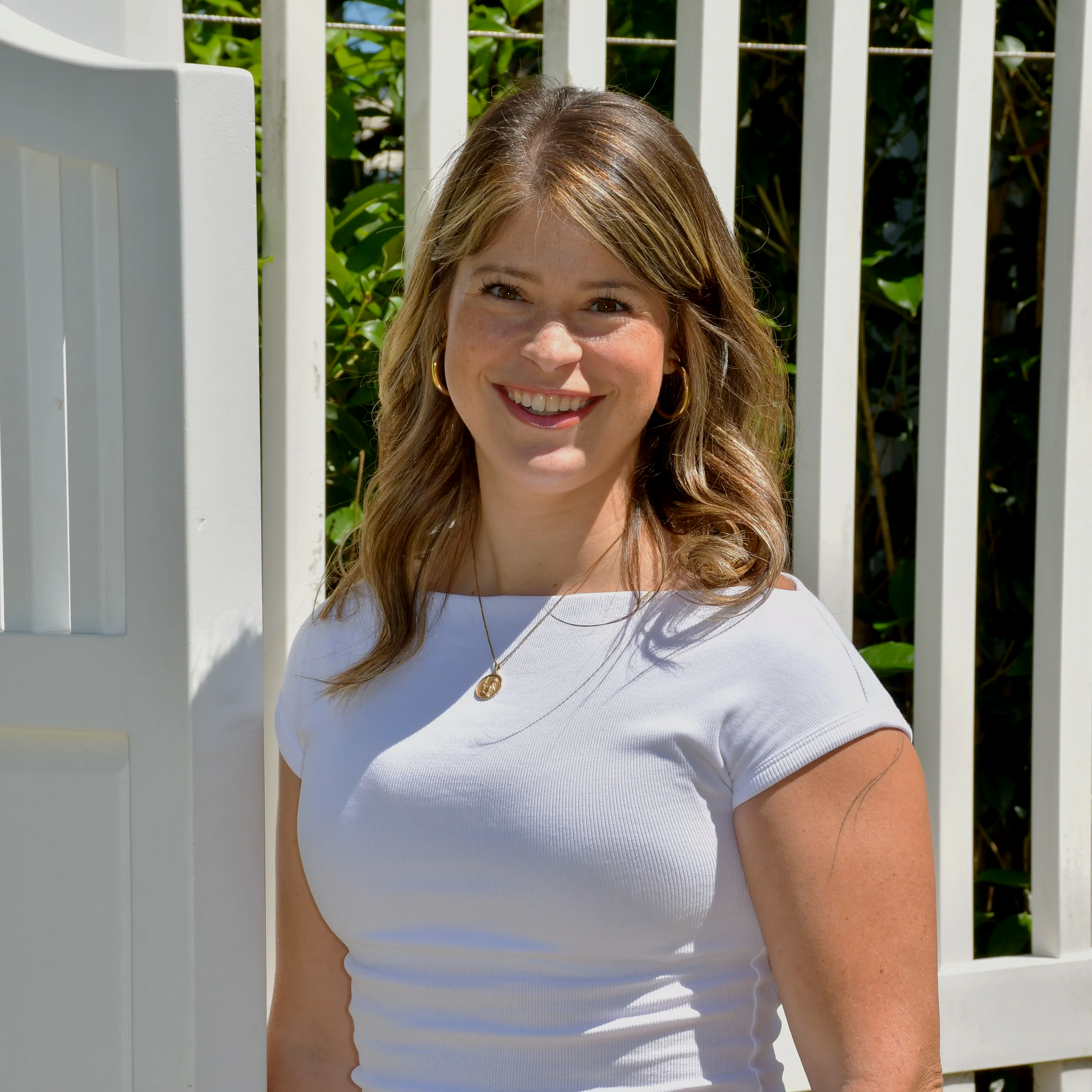 Close-up of a smiling young woman with light brown hair wearing a white top and gold jewelry, standing outdoors in front of a white picket fence with green foliage in the background.