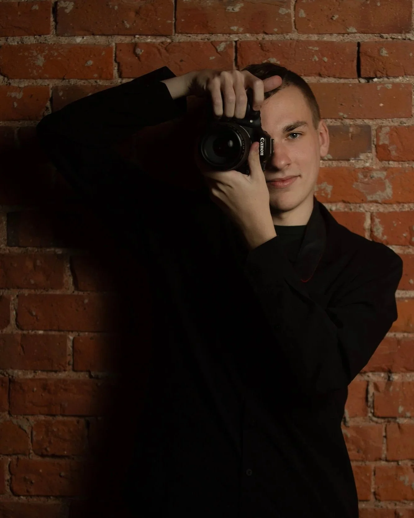 A young man in a black shirt taking a selfie with a Canon camera in front of a red brick wall.
